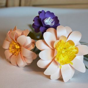 Bouquet de trois fleurs en papier blanc avec étamines jaunes et feuillage vert, sur socle arrondi.