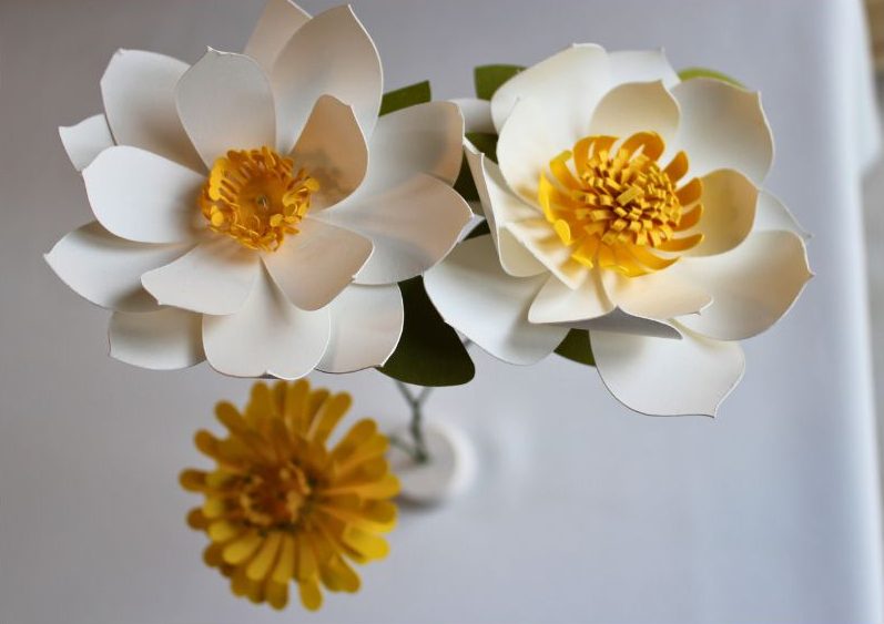 Bouquet de trois fleurs en papier blanc avec étamines jaunes et feuillage vert, sur socle arrondi.